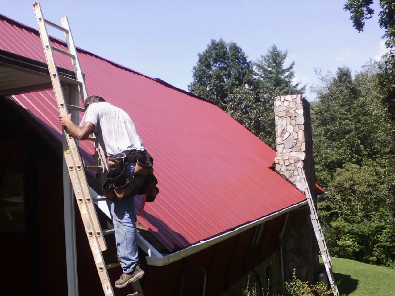 Roofer on a ladder beside a red metal roof and stone chimney — Soffit & Fascia Repair by Roofing Brothers in Zionsville